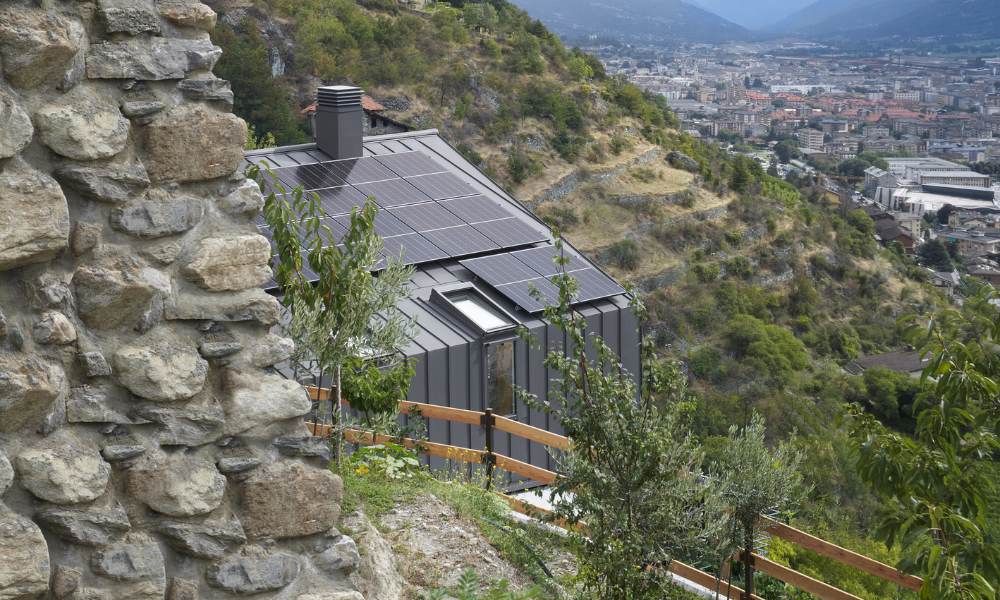 Casa moderna su un pendio montano, con tetto in metallo scuro e linee minimaliste, affacciata su una valle verde con un piccolo paese e montagne sullo sfondo sotto un cielo parzialmente nuvoloso.