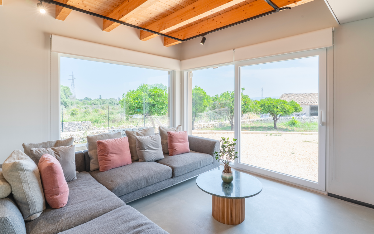 Un particolare della zona living, con il grande divano con il grigio e la sfumatura salmone che riprendono la tinta d'accento alla parete dell'ingresso e il microcemento del pavimento. La vista spazia indisturbata con i grandi infissi in pvc posizionati ad angolo.